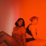 Vibrant studio portrait celebrating body positivity with two plus-size women sitting back to back.